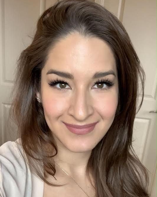 Woman with long brown hair wearing a beige top indoors.